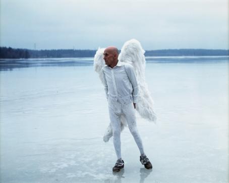 Vanha mies valkoisissa vaatteissa ja valkoiset enkelinsiivet selässään seisoo järven jäällä sinisävyisessä maisemassa.