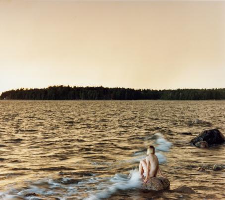 Aallokko, jonka taustalla näkyy metsää. Kivellä istuu alaston ihminen polvet koukussa, aalto lyö hänen jalkoihinsa.