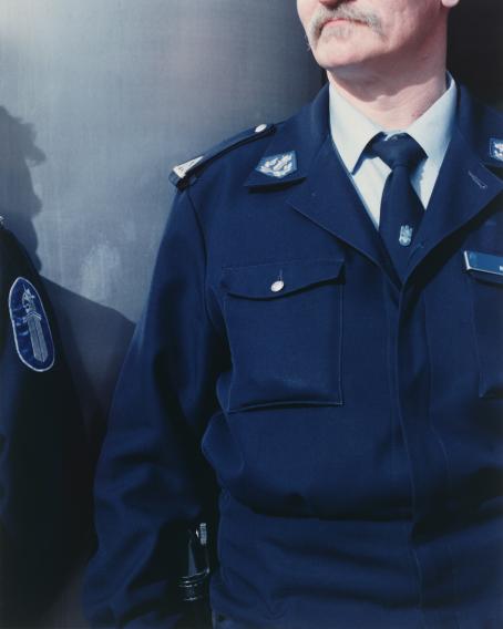 Poliisi, josta näkyy vain yläruumis. Kasvoista näkyvät suu ja vähän nenää. Vieressä on toinen poliisi, josta näkyy vain hihassa oleva poliisin tunnus.
