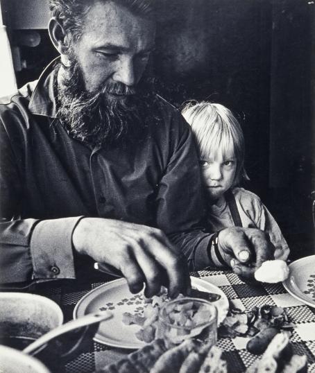”Mitään ruusuilla tanssimista Frimanien perheen elämä ei ollut. Mutta vaikka he uurastivat aikaisesta aamusta myöhäiseen iltaan, he olivat silti iloisia ja puheliaita. Frimaneja en unohda ensi hätään.” Kuvatakseen pienviljelijäperheen arkea Muoniossa vuonna 1968 Caj Bremer muutti viikoksi asumaan Frimanin perheen luo. Viisilapsinen perhe oli joutunut muuttamaan ja aloittamaan elämänsä alusta saadakseen riittävästi viljelysmaata. Kuva: Caj Bremer - Friman, uudisraivaaja, 1973 Mustavalkoisessa kuvassa parrakas mies istuu ruokapöydässä ja kuorii perunaa. Miehen vieressä istuu lapsi ja katsoo ujosti suoraan kameraan miehen takaa.