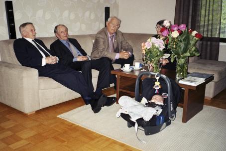 Sohvalla istuu kolme vanhaa miestä ja yksi vanha nainen. Sohvapöydällä on kahvikuppeja ja lautasia ja maljakoissa kukkia. Nainen jää osittain kukkien taakse. Pöydän edessä lattialla nukkuu vauva turvakaukalossa. 